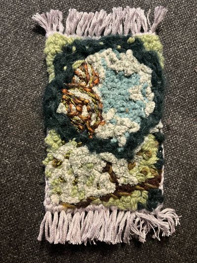 A small fringed textile mat with punch-needle embroidery depicting a mossy forest scene: pale blue-white clouds or stones, a gold and orange mushroom cluster, light green moss, and tiny gold bees, bordered in dark green.