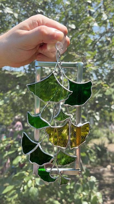 A rectangular metal frame with cascading green and gold stained-glass ginkgo leaves suspended on wire branches, photographed outdoors against foliage.