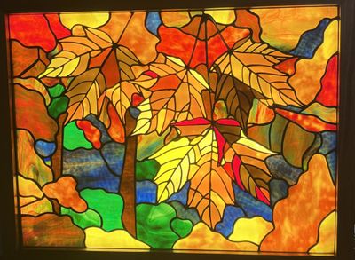 A backlit stained-glass panel in a wood frame depicting overlapping maple leaves in red, orange, and yellow, with tree trunks and a blue stream in the background.