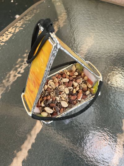 A triangular stained-glass birdfeeder in swirled amber and orange glass, hung by a black leather cord and filled with mixed birdseed and nuts.