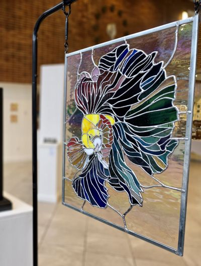 A large rectangular stained-glass panel of a betta fish with iridescent green, blue, and jewel-toned fins fanning outward, hung on a metal frame stand.