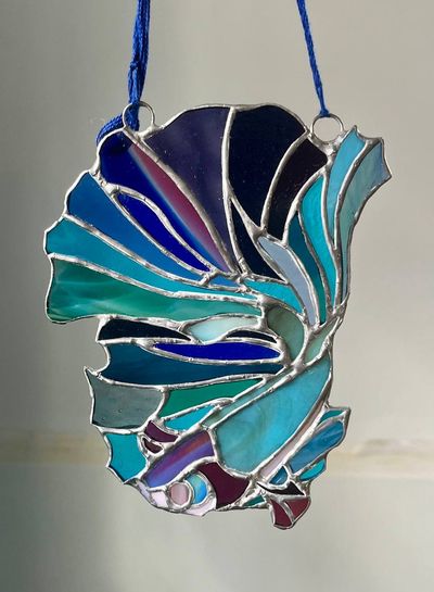 A stained-glass betta suncatcher with flowing blue, teal, and violet fins curving back on itself, hanging from blue cords.