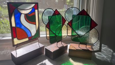 Three stained-glass trophies on wood bases: an abstract panel of curving red, blue, cream, and green shapes, and two faceted pieces with layered green glass, red triangle points, and a textured clear panel etched with a baseball's stitching.