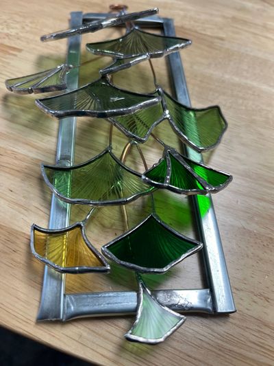 A rectangular metal frame with layered green and amber stained-glass ginkgo leaves cascading down copper wire branches.