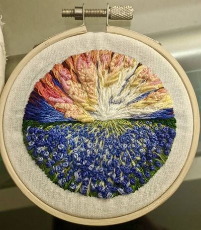 A round embroidery hoop piece showing a radiating sunset sky in pink, orange, yellow, and blue thread above a field of blue bonnet flowers in blue and green.