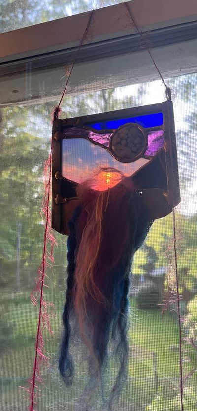 A small stained-glass panel depicting a sunset sky in blue, purple, and orange with an agate stone inset, hung in a window with cascading strands of blue, orange, and pink wool and silk roving flowing down beneath it like a waterfall.