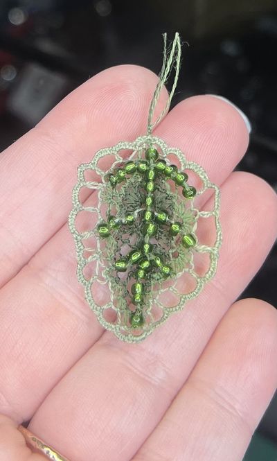 A small oval leaf shape in sage-green needle lace with a scalloped crocheted border and a center vein of dark green glass beads.