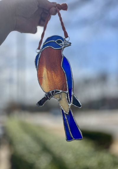 A stained-glass bluebird with a blue head, wings, and tail, a rust-orange breast, and a cream belly, perched on a copper wire branch and hung from a cotton cord.