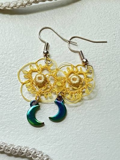 A pair of dangle earrings with yellow crocheted lace sunflower motifs and iridescent teal-blue crescent moon charms, on silver ear wires.