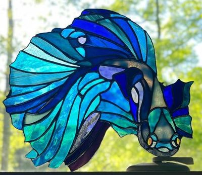 A stained-glass betta fish panel with sweeping blue and teal fins, mounted upright and photographed head-on against a wooded outdoor view.