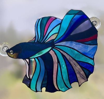 A stained-glass betta fish suncatcher in deep blues, teals, and magenta with sweeping fan-shaped fins, mounted with suction cups against a hazy outdoor backdrop.
