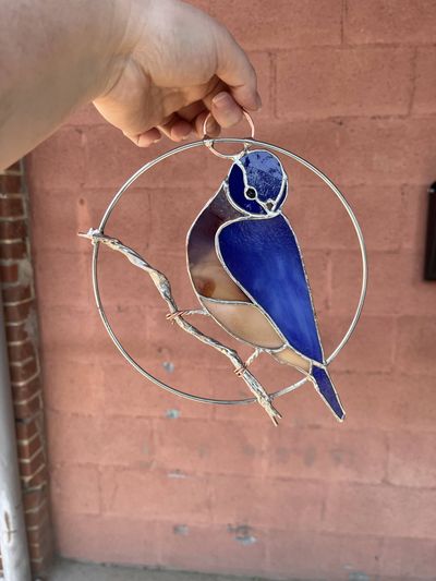 A stained-glass bluebird with a royal blue head, wings, and tail, and a tan belly, perched on a wire branch inside a circular metal hoop.