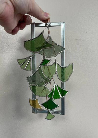 A rectangular metal frame with cascading green and gold stained-glass ginkgo leaves hanging from wire branches.