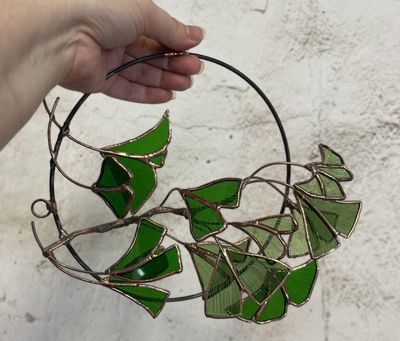 A circular wire hoop wrapped with branching copper wire and clusters of green stained-glass ginkgo leaves in varying shades, arranged in a spiral around the ring.
